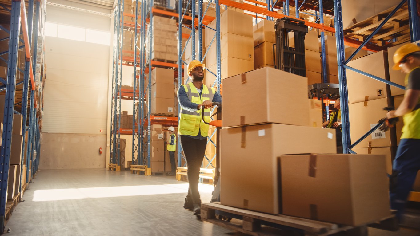 Middle mile logistics is concerned with moving products through the supply chain's intermediary steps shown with diverse workers in a warehouse moving product from the production floor to warehouse— in preparation for last-mile shipping.