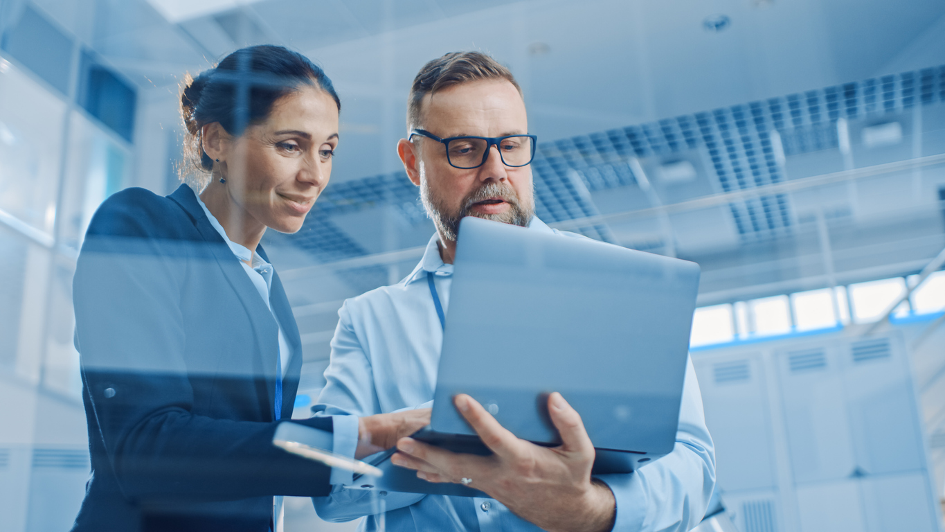 Businesswoman and man working on a supply chain branding and marketing, man holds laptop computer, they reference it. In the Background Modern and Bright Facility. Low Angle Shot