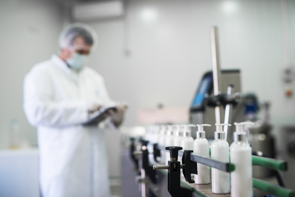 Close-up of liquid soap on a cosmetics production line in front of a worker typing on a tablet in a cGMP-compliant warehouse.