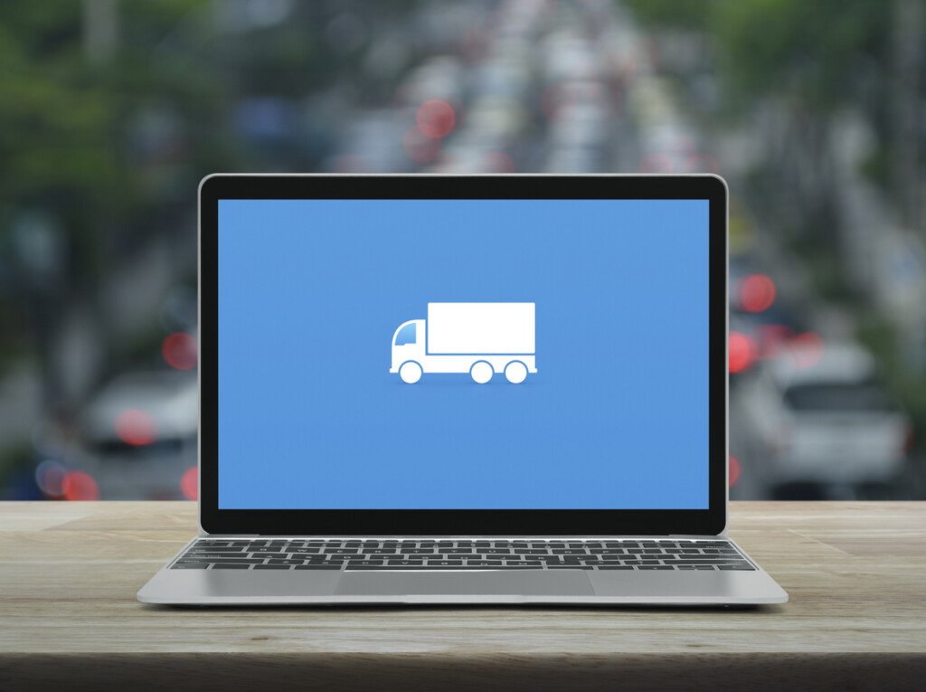 Delivery truck flat icon on modern laptop computer screen on wooden table over blur of rush hour with cars and road in city, Business transportation online concept