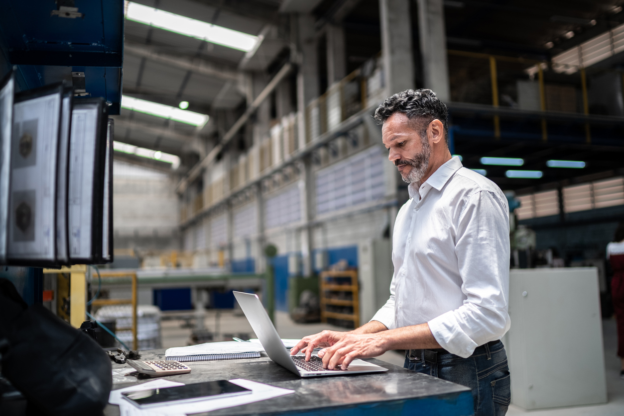 Mature businessman using a laptop in a modern logistics warehouse, highlighting technology and supply chain advancements.