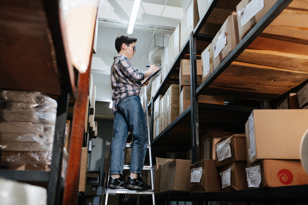 A female warehouse worker standing on a ladder amidst rows of shelves filled with boxes, reviewing a clipboard. This scene illustrates the process of identifying changing pain points in supply chain accounts.