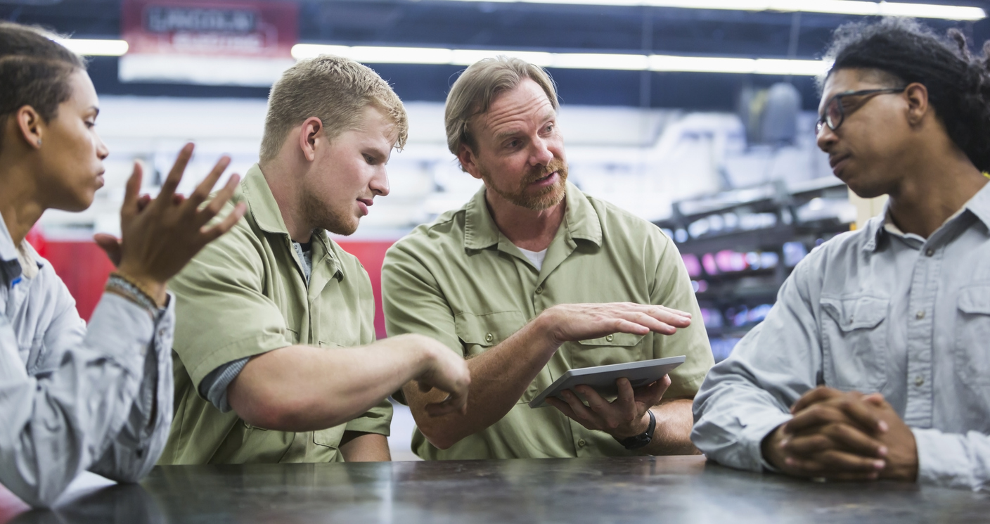 Four people discussing outsourcing their auto parts to a 3rd party logistics company. Man holding a digital tablet, talking about auto parts fulfillment and looking at the man sitting next to him.