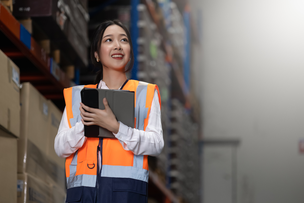 Professional woman in an orange vest using a digital tablet to check stock and inventory in an efficiently organized warehouse or a dedicated facility in action. Illustrating the role of dedicated warehousing in logistics and distribution center operations.