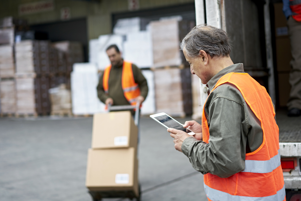 Focus on foreground man in casual clothing and reflective vest checking portable information device as coworker moves boxes with hand truck in background.