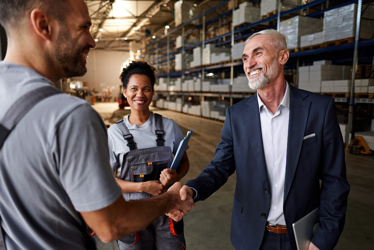 A cheerful manager and freight worker shake hands while a colleague looks on in a distribution warehouse, exemplifying strong supplier and freight relationships essential for cost reduction in logistics.