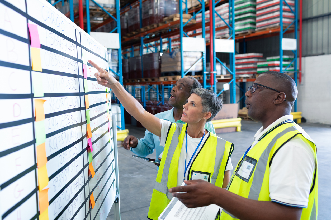Warehouse staff discussing plans and reviewing a whiteboard in a warehouse, emphasizing the importance of setting clear expectations in fulfillment partnerships