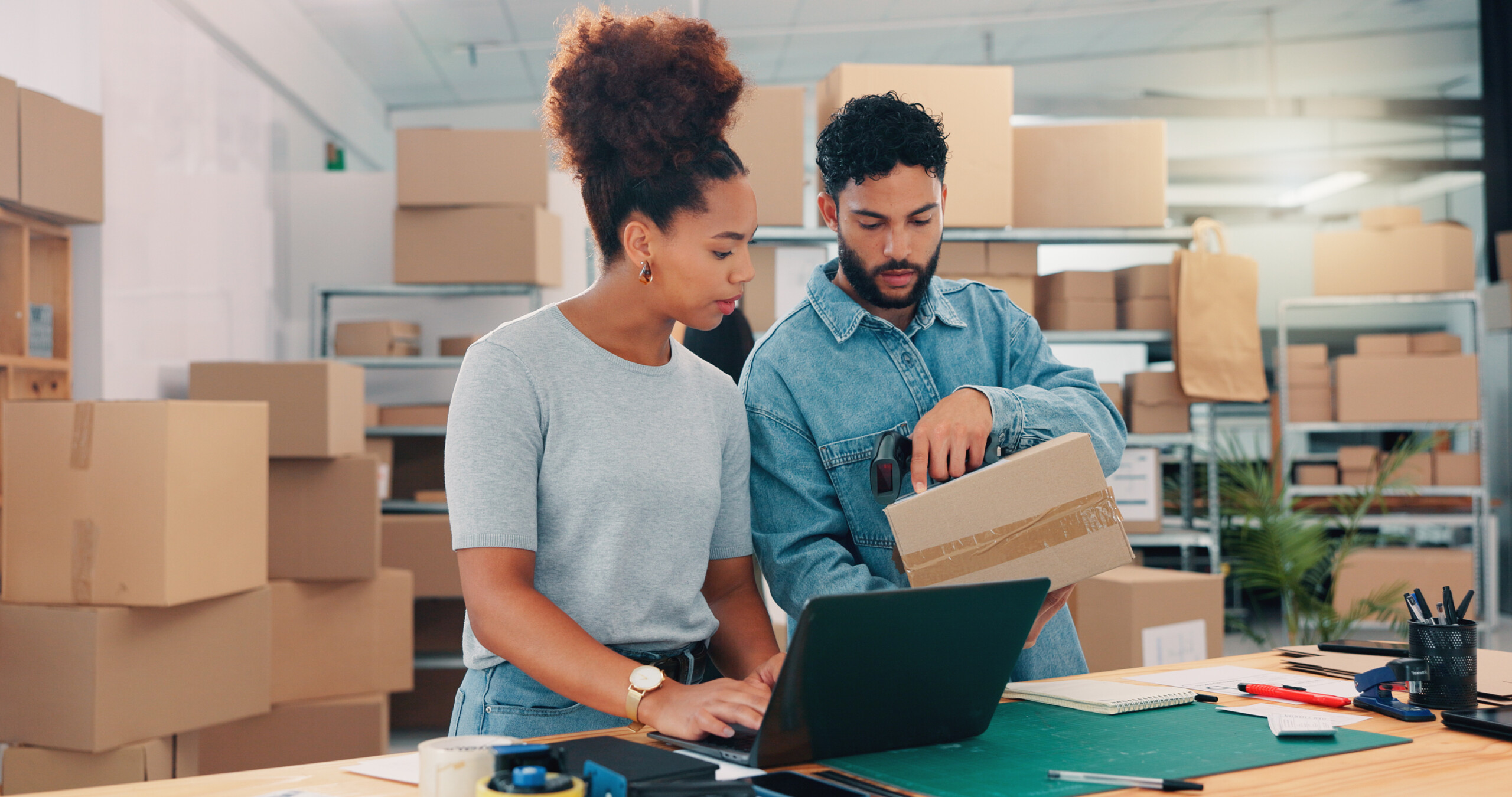 Small business owners reviewing order fulfillment data on laptop while handling shipping packages in modern fulfillment workspace